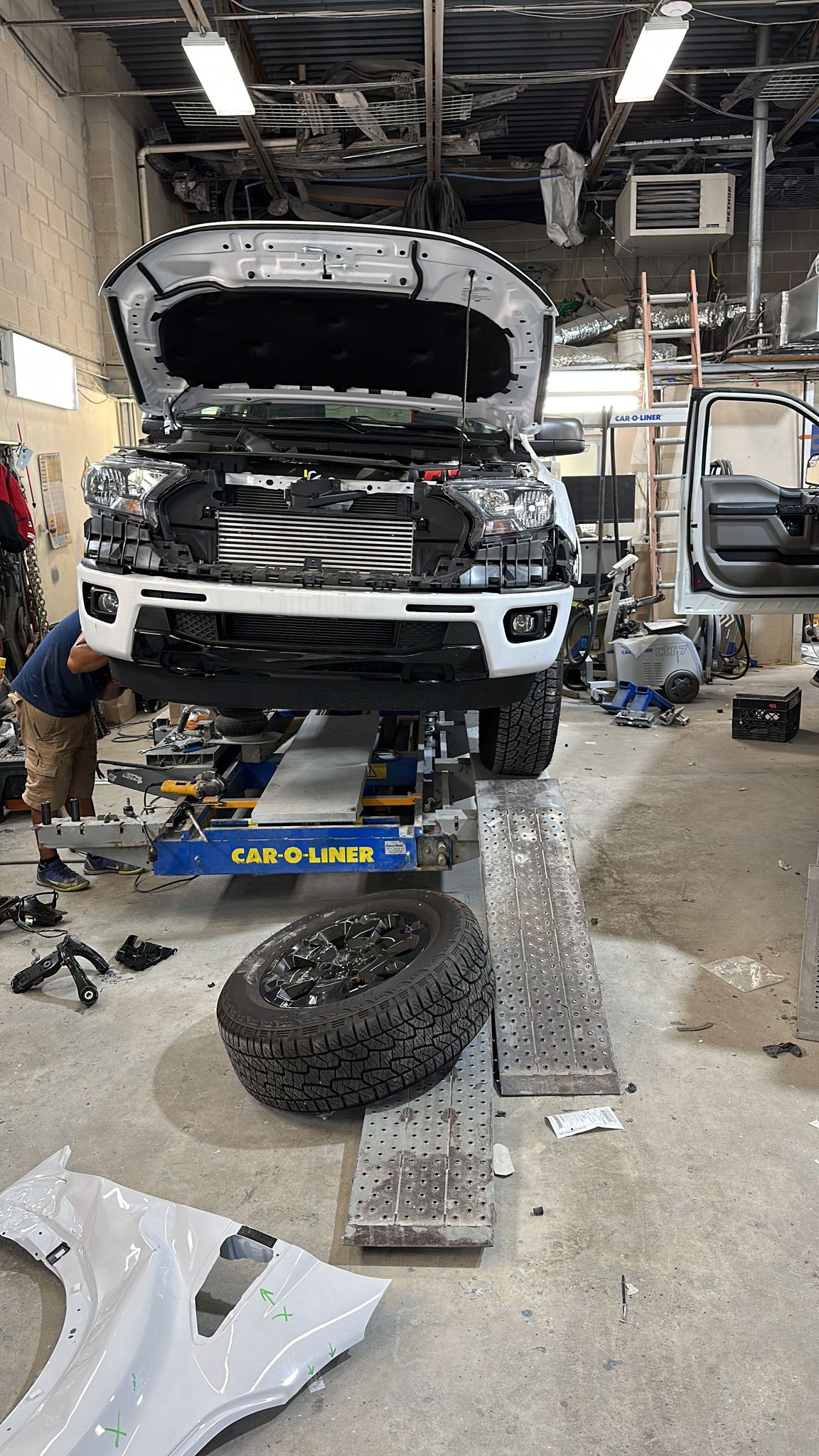 White pickup with its front bumper removed on a frame bench inside the shop