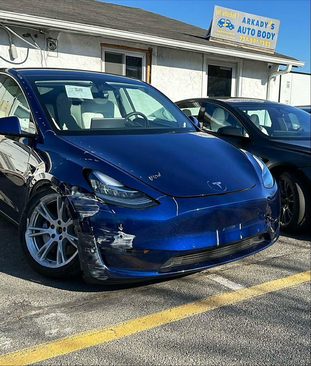 Blue Tesla with front-corner damage parked below Arkady's shop sign